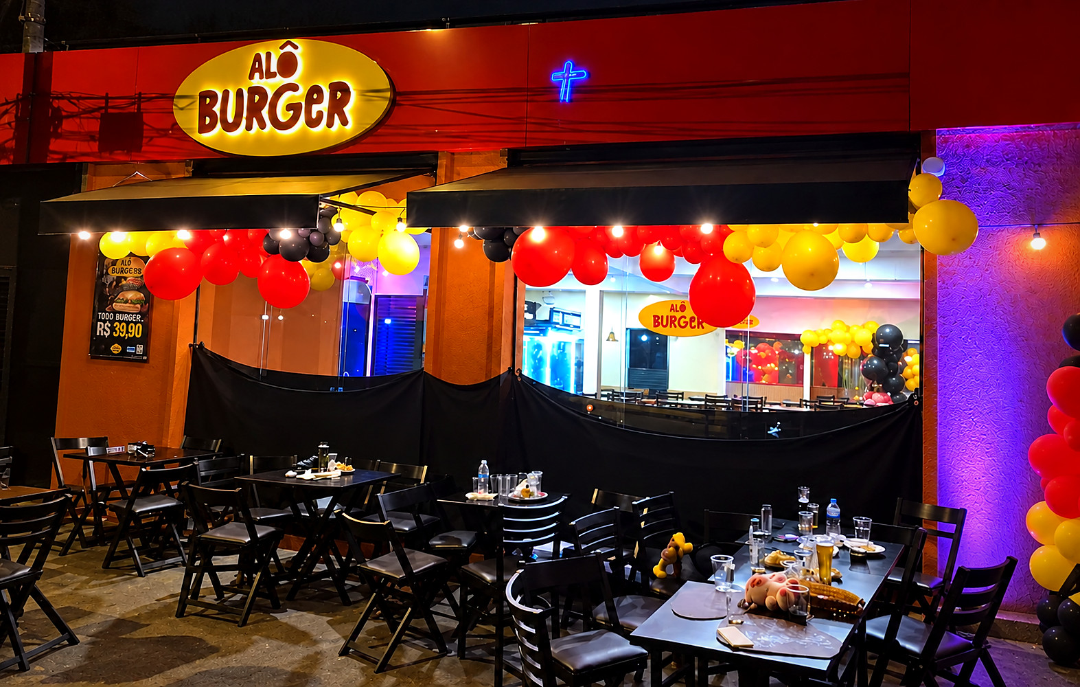 Fachada iluminada da Alô Burger, com mesas e decoração em amarelo, vermelho e preto
